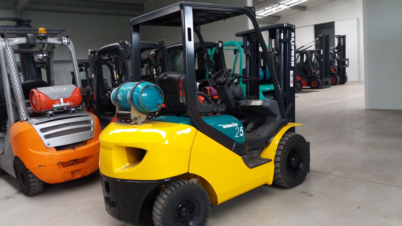 Żółto-czarny wózek widłowy Komatsu 25 z niebieską butlą gazową na tle magazynu z innymi wózkami widłowymi.