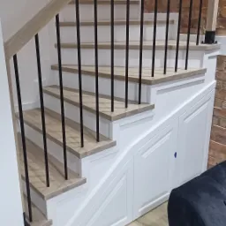 Białe schody z drewnianymi stopniami i czarną metalową balustradą, zintegrowane z szafkami wnękowymi, na tle ściany z cegły.