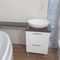 Jasna łazienka na poddaszu ze skośnym sufitem, z białą umywalką nablatową na szafce z dwiema szufladami, obok toaleta z dwoma rolkami papieru na górze, wanna oraz drewniany blat w kolorze ciemnego...