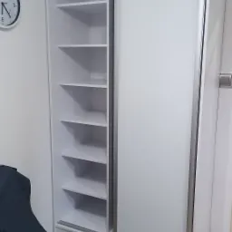 Biała szafa przesuwna z półkami, jedna strona otwarta, widoczny zegar na ścianie i fragment łóżka z ciemną narzutą.