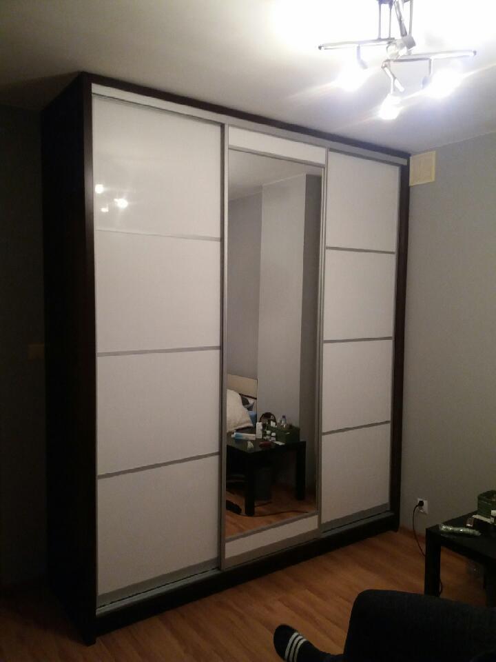 Duża szafa przesuwna z białymi frontami i lustrem na środkowych drzwiach, ciemnobrązowa rama, widoczny fragment pokoju z parkietem i stolikiem nocnym odbijającym się w lustrze.