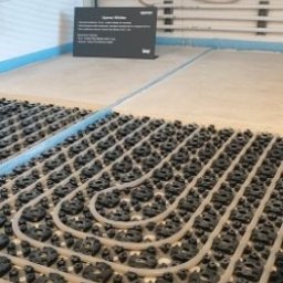 DB MONTER Dariusz Szczepaniak - Ułożona instalacja ogrzewania podłogowego z rurami w czarnych uchwytach, widok z góry, fragment ściany z instalacją w tle. Tabliczka informacyjna w lewym górnym rogu.