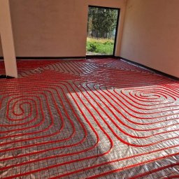 DB MONTER Dariusz Szczepaniak - Czerwone rury ogrzewania podłogowego ułożone spiralnie na folii izolacyjnej w surowym pomieszczeniu z widokiem na zieleń przez okno.