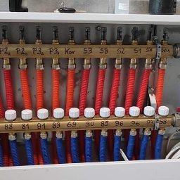 DB MONTER Dariusz Szczepaniak - Rozdzielacz ogrzewania podłogowego w białej szafce, z rurami w kolorach czerwonym i niebieskim, z widocznymi oznaczeniami na listwach rozdzielczych.