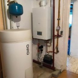 DB MONTER Dariusz Szczepaniak - Biały piec gazowy wiszący na ścianie, obok zbiornik na wodę OSO i instalacja z rur miedzianych. Widoczne elementy instalacji w piwnicy.