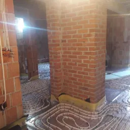 Wnętrze w trakcie montażu ogrzewania podłogowego, widoczne rury grzewcze ułożone na folii izolacyjnej, murowane ściany z czerwonej cegły w surowym stanie, wystające przewody elektryczne.