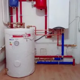 Nowoczesny, biały piec gazowy wiszący na ścianie z czerwonymi i niebieskimi rurami, połączony z cylindrycznym zbiornikiem na wodę, widoczne manometry i etykiety energetyczne, instalacja w kotłowni...