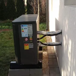 Zewnętrzna jednostka pompy ciepła Kaisai zamontowana na betonowych bloczkach przy ścianie budynku, rury zabezpieczone folią aluminiową, widoczne etykiety energetyczne i ostrzegawcze.