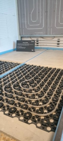 Ułożona instalacja ogrzewania podłogowego z rurami w czarnych uchwytach, widok z góry, fragment ściany z instalacją w tle. Tabliczka informacyjna w lewym górnym rogu.