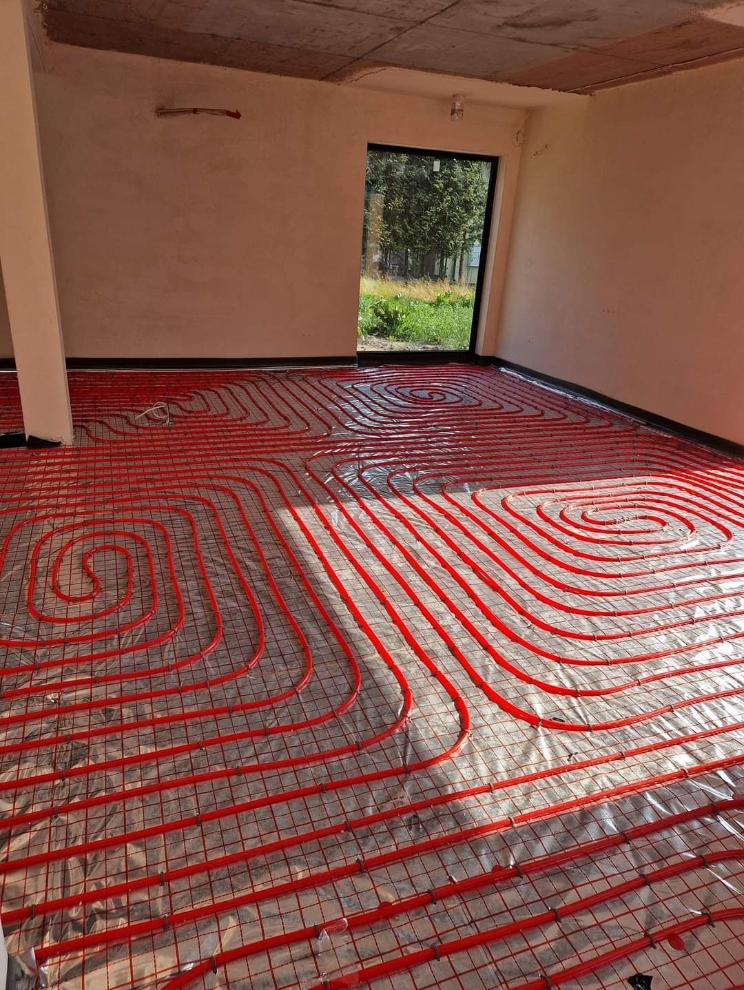 Czerwone rury ogrzewania podłogowego ułożone spiralnie na folii izolacyjnej w surowym pomieszczeniu z widokiem na zieleń przez okno.