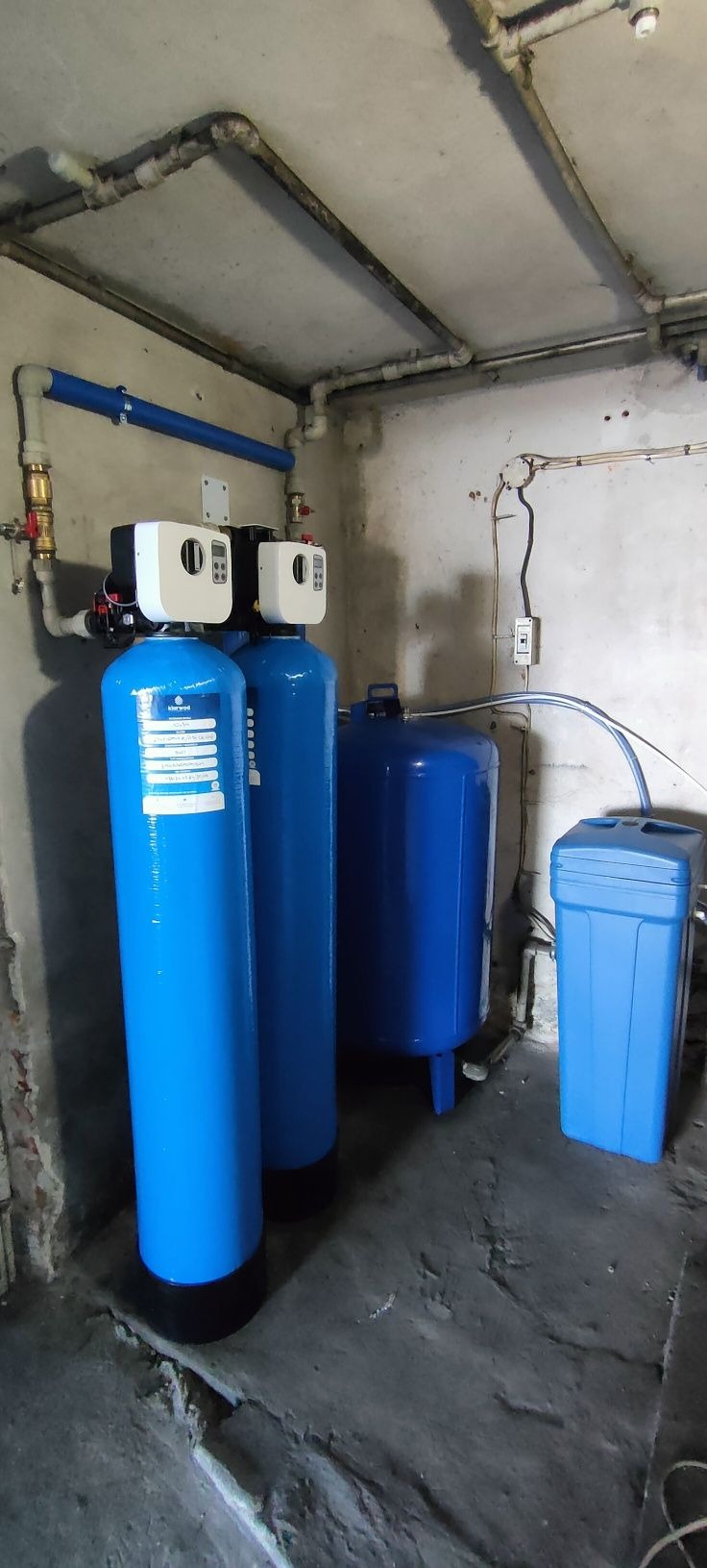 Niebieskie filtry do wody w piwnicy z rurami na suficie. Dwa filtry cylindryczne z białymi głowicami, obok zbiornik i pojemnik. Szare ściany i podłoga.