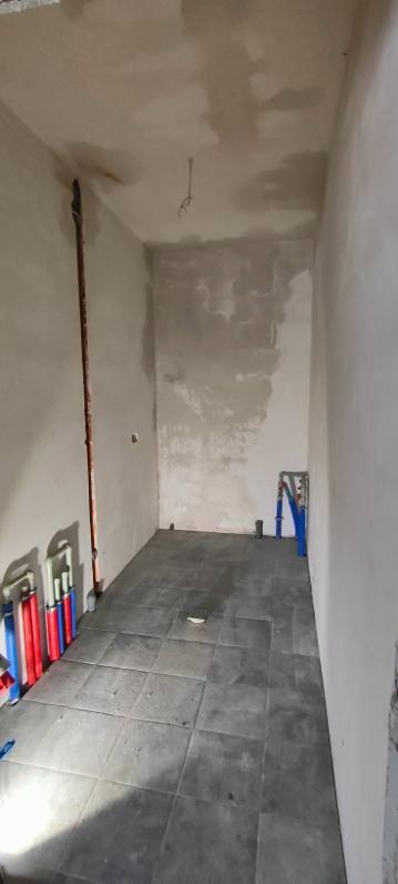 Wnętrze w trakcie remontu: szare płytki podłogowe, fragment instalacji hydraulicznej z rurami PEX w kolorach niebieskim i czerwonym, surowe ściany przygotowane do wykończenia.