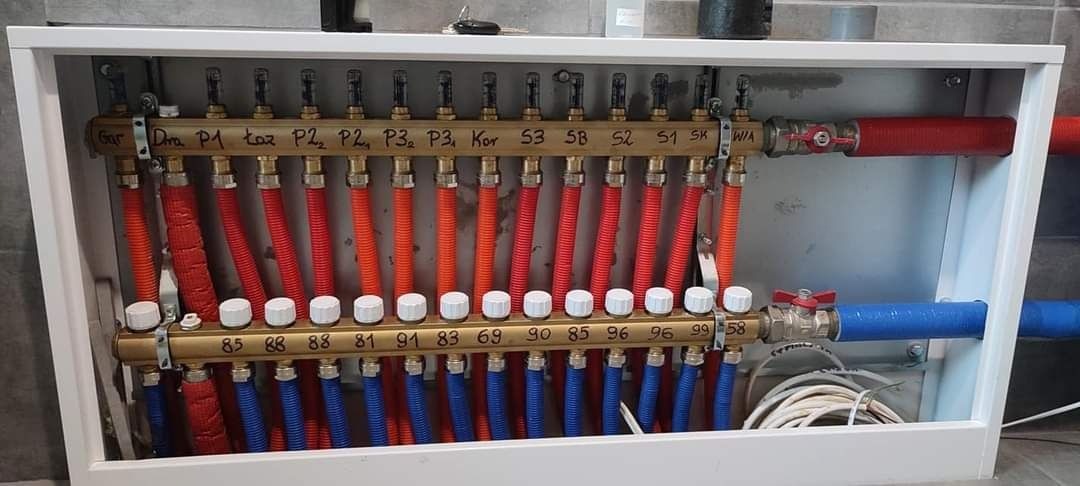 Rozdzielacz ogrzewania podłogowego w białej szafce, z rurami w kolorach czerwonym i niebieskim, z widocznymi oznaczeniami na listwach rozdzielczych.