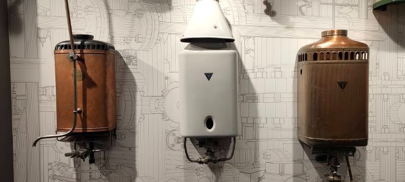 Trzy zabytkowe piece gazowe różnych modeli, umieszczone na ścianie z tapetą w techniczne rysunki. Jeden biały, dwa miedziane, prezentowane w muzeum.