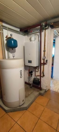 Biały piec gazowy wiszący na ścianie, obok zbiornik na wodę OSO i instalacja z rur miedzianych. Widoczne elementy instalacji w piwnicy.