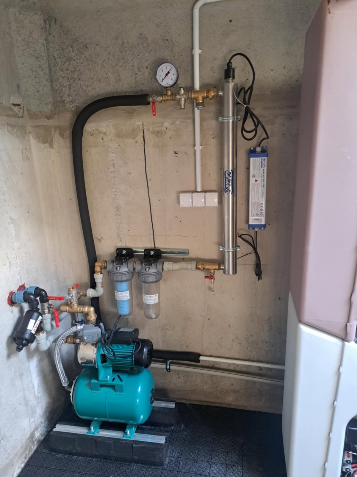 Zestaw hydroforowy z filtrami i lampą UV, zainstalowany w pomieszczeniu o surowych, betonowych ścianach. Widoczny manometr i rury doprowadzające wodę.