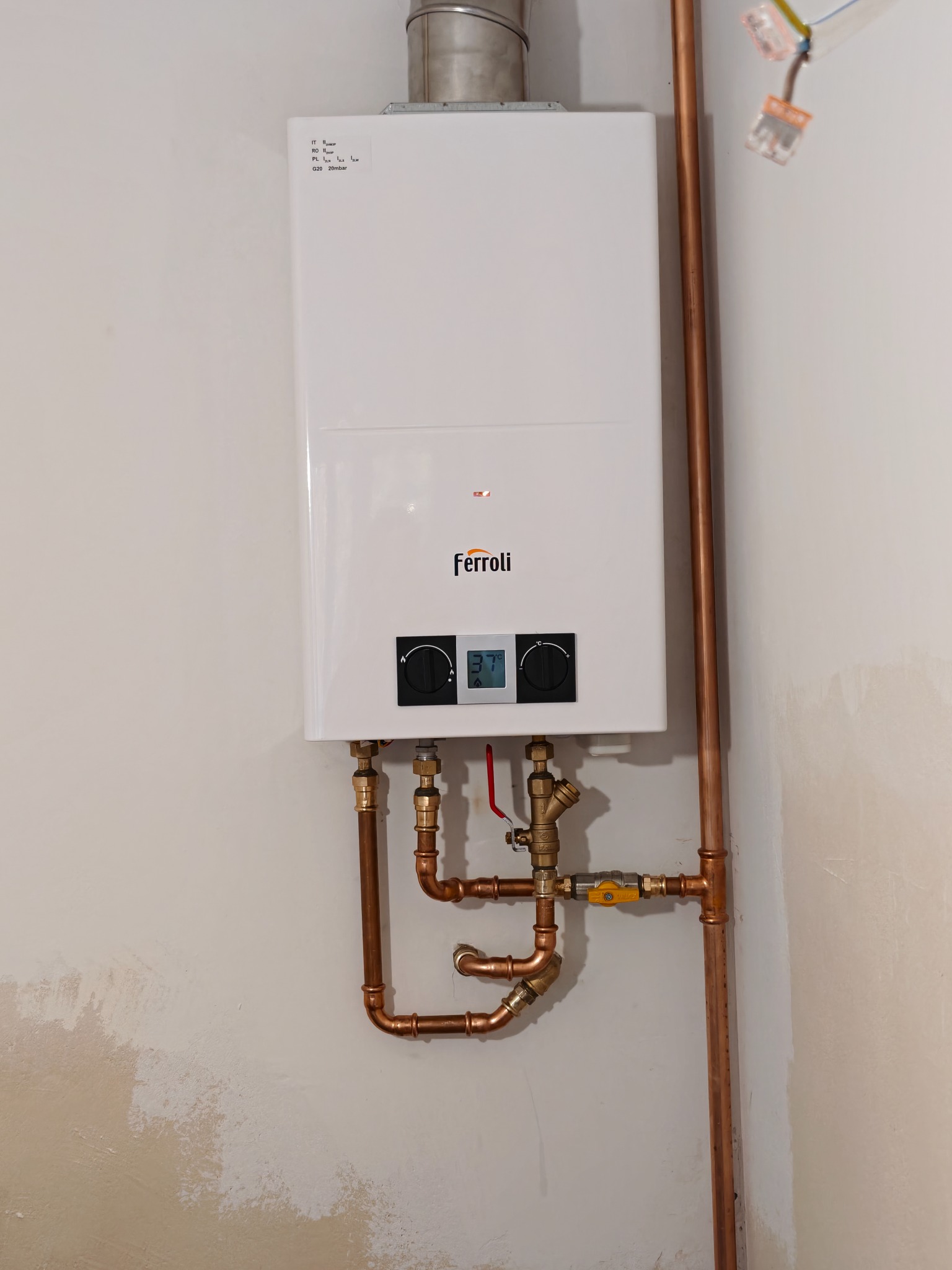 Biały piec gazowy Ferroli zamontowany na ścianie z miedzianymi rurami. Widoczne przyłącza i zawory. Minimalistyczny design pieca.