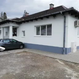 Biały budynek z szarą podmurówką i ciemnym dachem, widoczny klimatyzator na elewacji, szary samochód zaparkowany obok, na pierwszym planie teren utwardzony kostką brukową i gruzem.