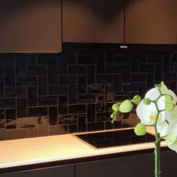Nowoczesna kuchnia z czarnymi, błyszczącymi płytkami ułożonymi w cegiełkę, jasnym blatem i białymi gniazdkami elektrycznymi, ozdobiona białą orchideą.