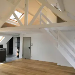 Jasne poddasze z drewnianą podłogą, białymi belkami stropowymi, schodami z białymi poręczami i częściowo widoczną kuchnią z czarnymi blatami.