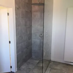 Nowoczesna kabina prysznicowa typu walk-in z szarymi płytkami imitującymi beton, deszczownicą sufitową i szklaną ścianką zabezpieczoną ż&oacute;łtymi podkładkami montażowymi.