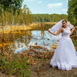 Panna młoda w białej sukni i welonie pozuje na tle jeziora z powalonymi drzewami i lasem w tle.