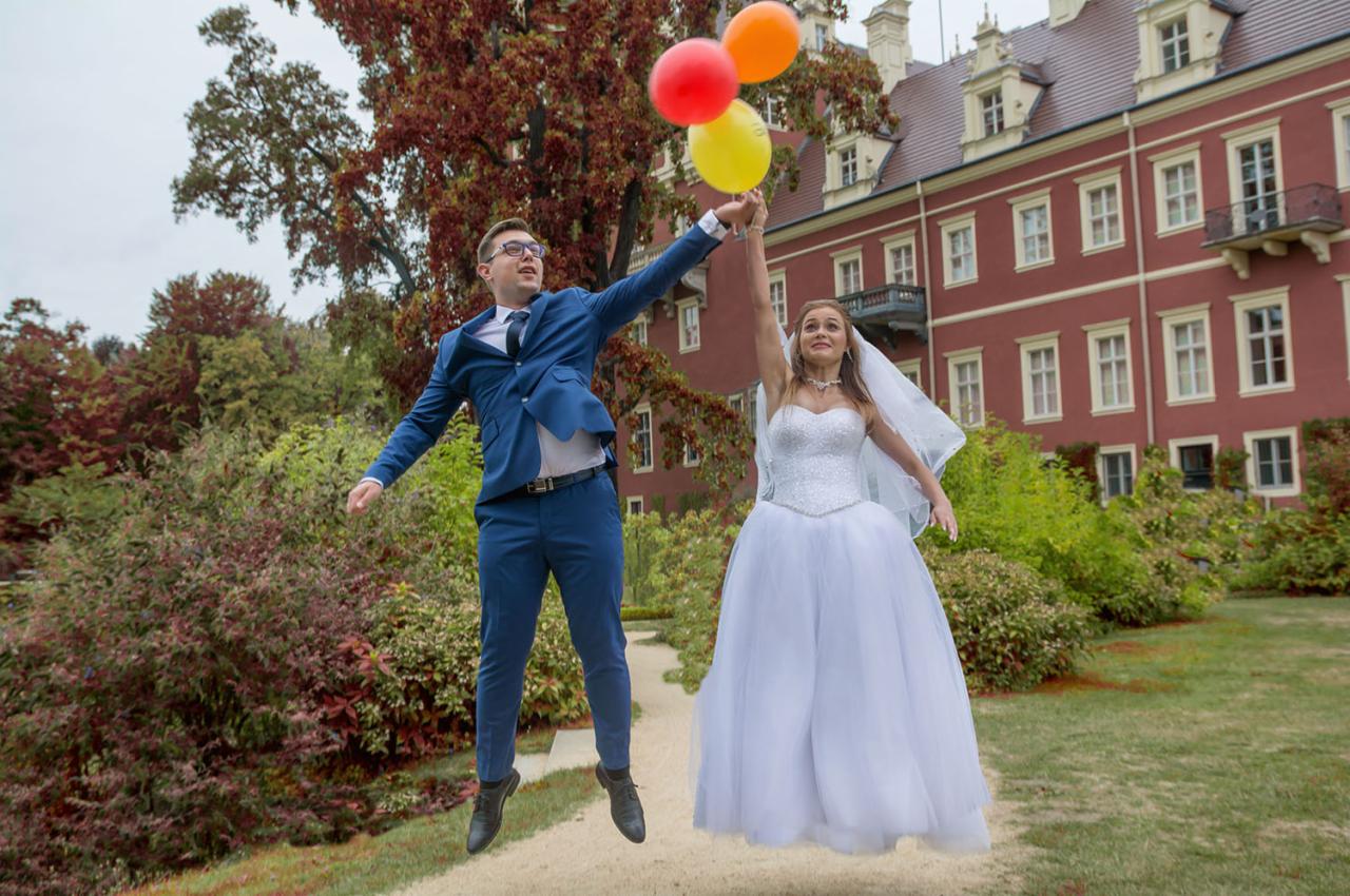 Para młoda w skoku radości z kolorowymi balonami na tle czerwonego budynku w parku.