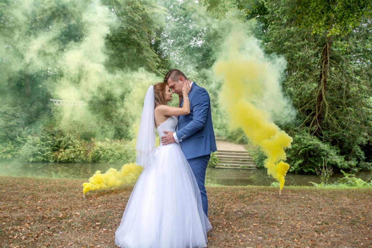 Para młoda w objęciach na tle żółtego i zielonego dymu w plenerze w Zielonej Górze, panna młoda w białej sukni i welonie, pan młody w niebieskim garniturze.