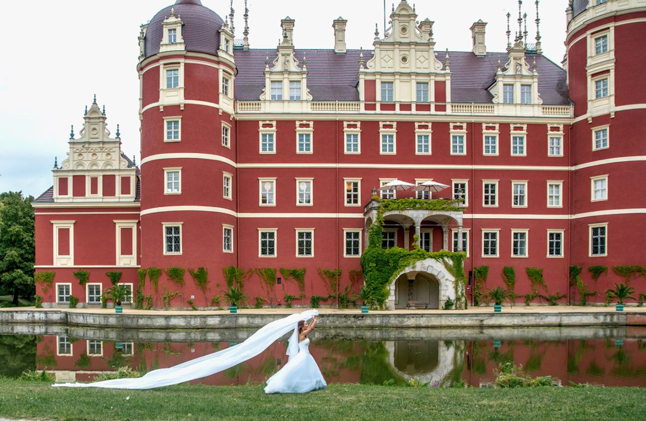 Panna młoda w białej sukni i długim welonie pozuje na tle czerwonego zamku odbijającego się w stawie.