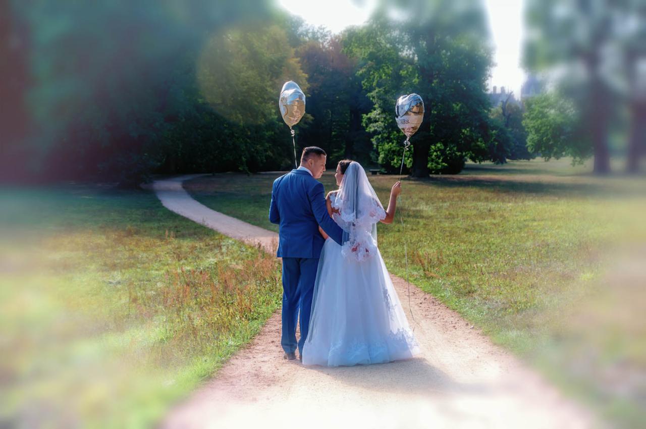 Para młoda w niebieskim garniturze i białej sukni spaceruje ścieżką w parku, trzymając balony w kształcie serc. Widok z tyłu, z rozmytym tłem drzew i łąki.