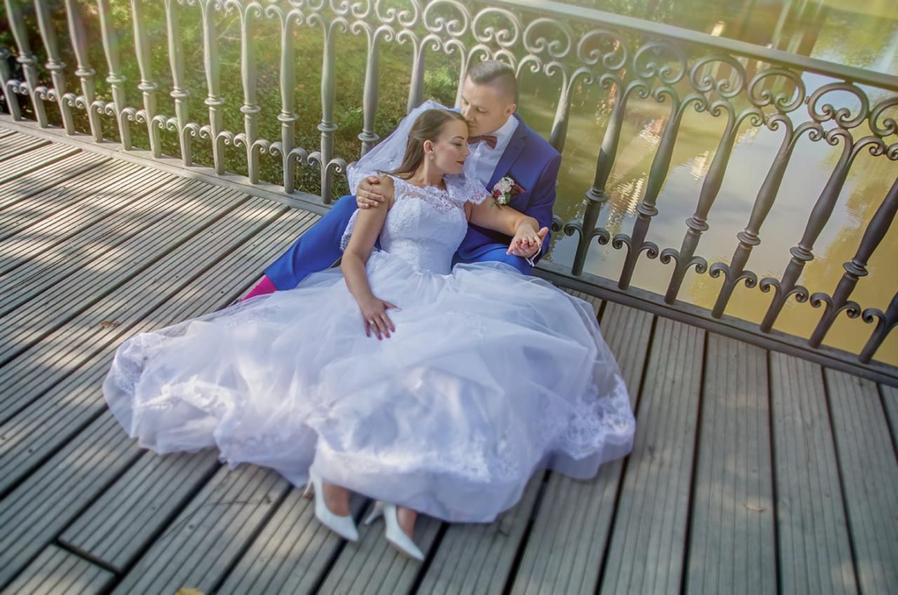 Para młoda siedzi na drewnianym moście, on w niebieskim garniturze z różowymi skarpetkami, ona w białej sukni ślubnej z koronkowymi detalami, w tle woda i zieleń.