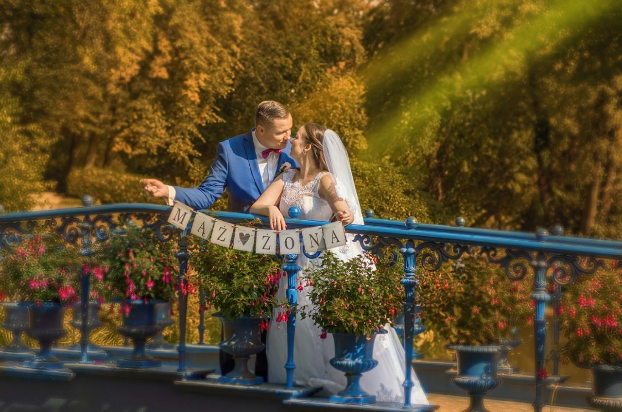 Para młoda, on w niebieskim garniturze z muszką, ona w białej sukni z welonem, pozuje na niebieskim moście z napisem 'Mąż i Żona', otoczonym donicami z kwiatami na tle jesiennego parku.