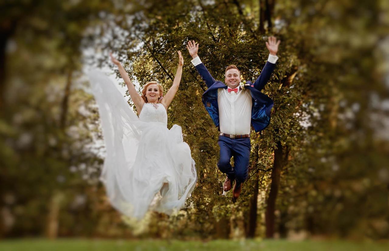 Para młoda w trakcie skoku radości na tle zieleni, panna młoda w białej sukni z rozłożystym dołem, pan młody w granatowym garniturze z muszką.
