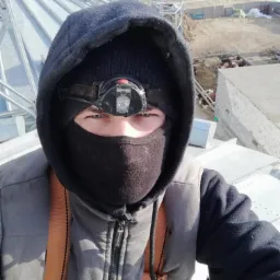 Pracownik w odzieży roboczej i z czołówką na głowie, zabezpieczony szelkami, stoi na konstrukcji silosu zbożowego, w tle zabudowania gospodarcze i pole.