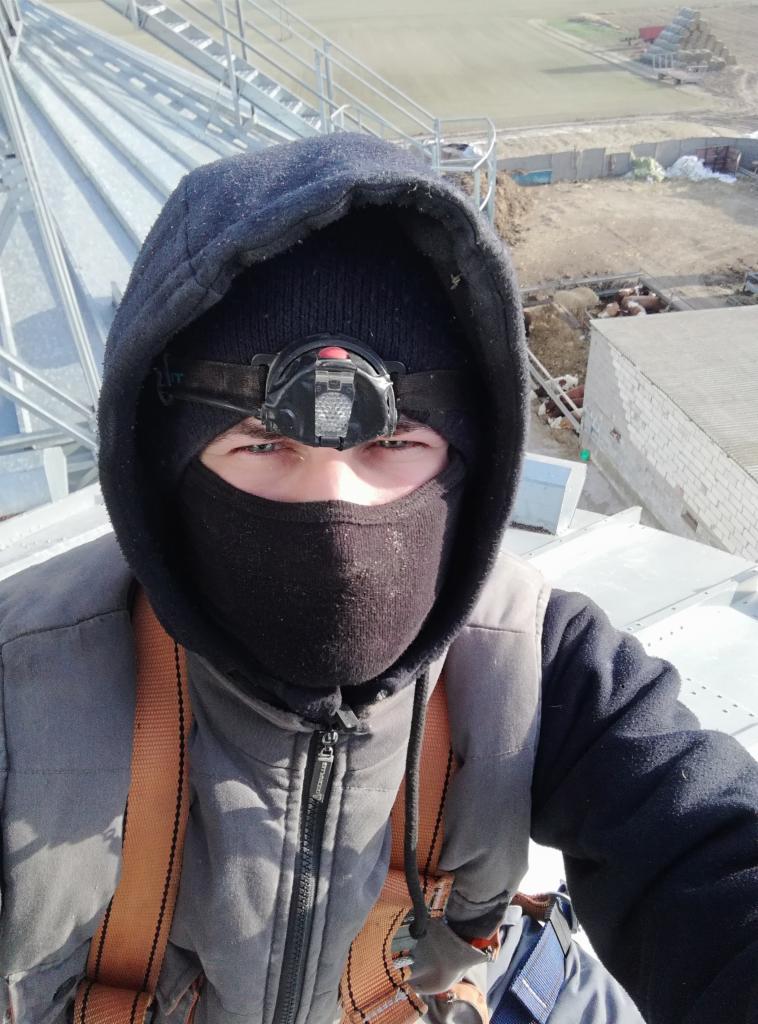 Pracownik w odzieży roboczej i z czołówką na głowie, zabezpieczony szelkami, stoi na konstrukcji silosu zbożowego, w tle zabudowania gospodarcze i pole.