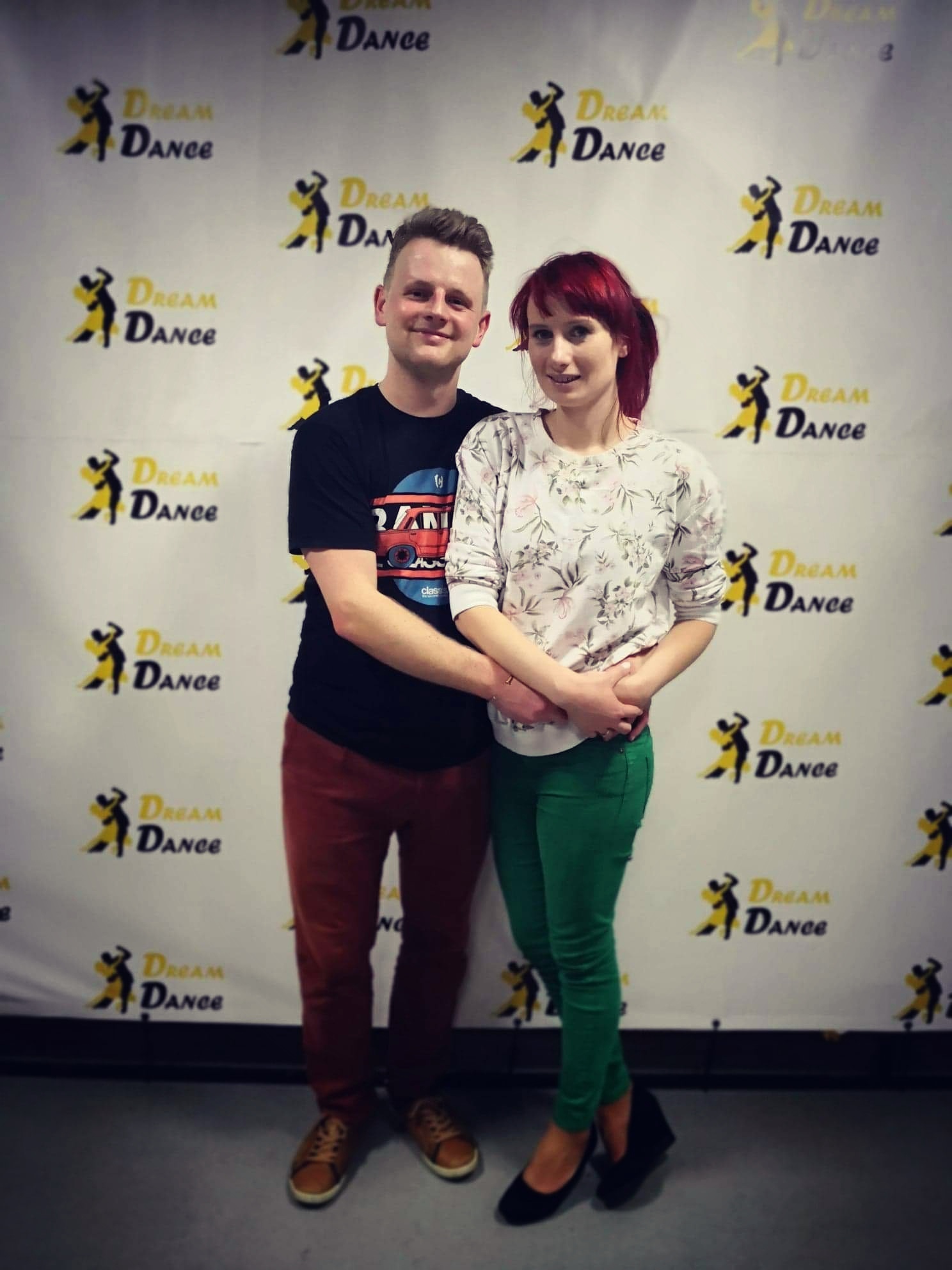 Para uśmiechniętych tancerzy pozuje na tle ściany z powtarzającym się logo 'Dream Dance' w kolorze żółto-czarnym.