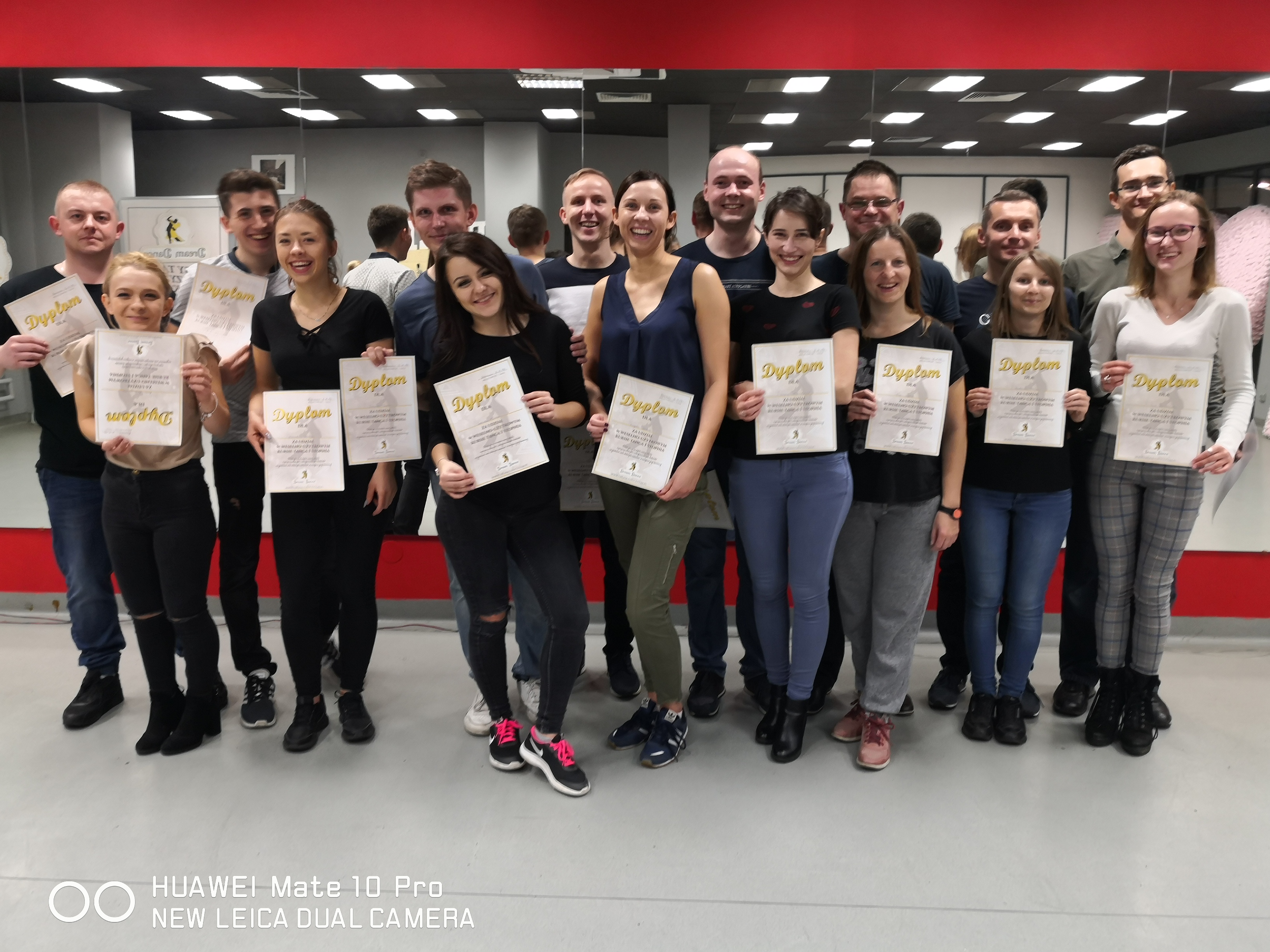 Grupa uśmiechniętych osób, prawdopodobnie kursantów, trzyma dyplomy ukończenia kursu na tle lustra w sali.