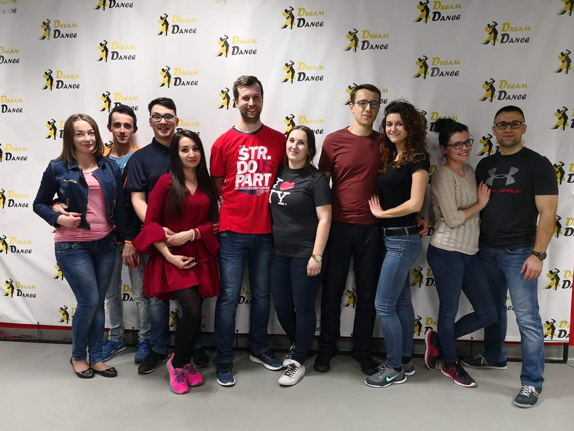 Grupa uśmiechniętych osób pozuje na tle ściany z powtarzającym się logo 'Dream Dance', widać różnorodne stroje, od casualowych jeansów po elegancką czerwoną sukienkę.