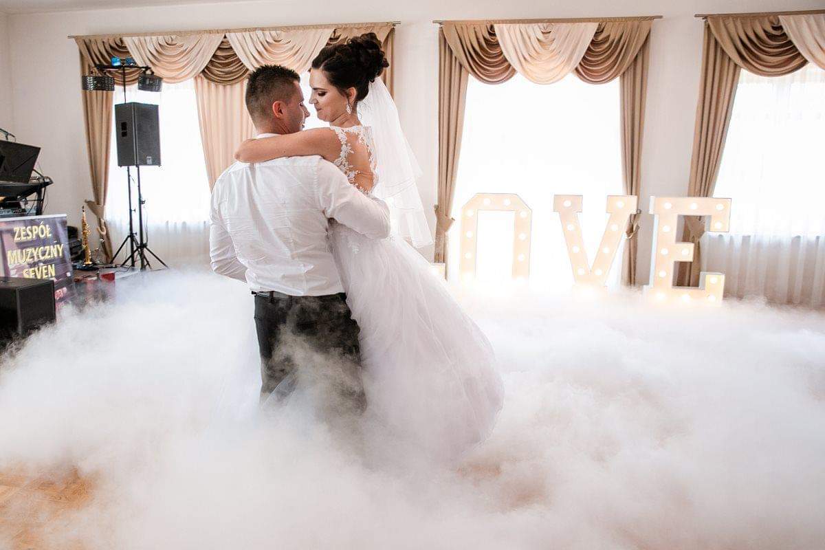 Para młoda tańczy pierwszy taniec w chmurze dymu, dekoracja LOVE z podświetleniem w tle, zespół muzyczny widoczny w lewym rogu.
