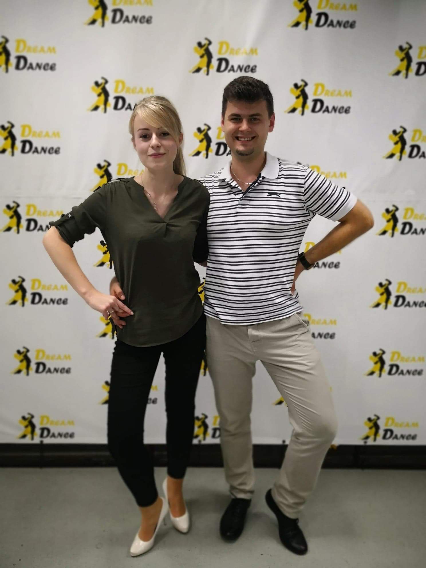 Instruktorzy tańca pozują na tle ściany z powtarzającym się logo i nazwą firmy, ona w czarnych spodniach i oliwkowej bluzce, on w beżowych spodniach i koszulce w paski.