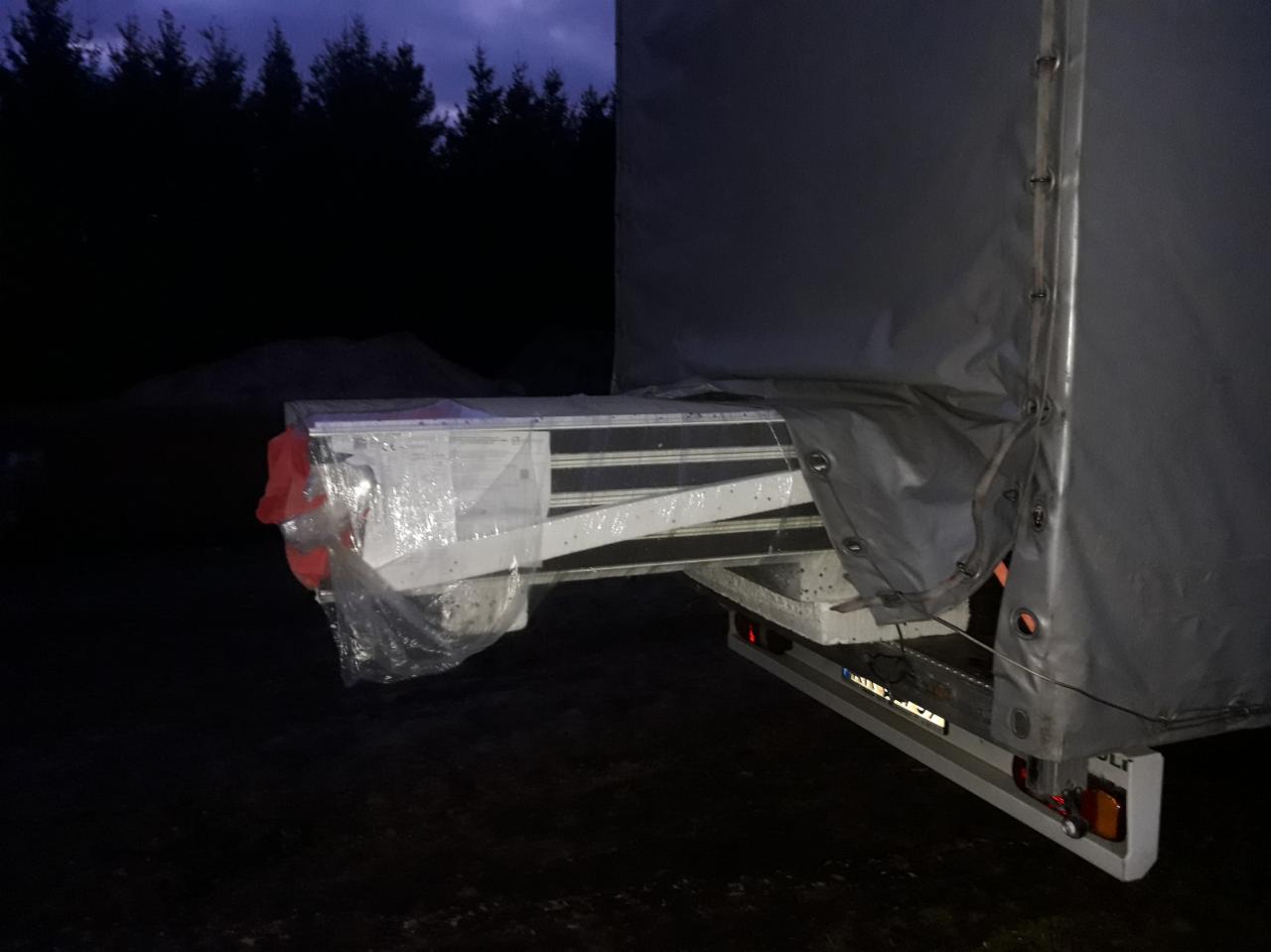Fragment naczepy z ładunkiem zabezpieczonym folią, widoczne tylne światła i plandeka, w tle ciemny las.