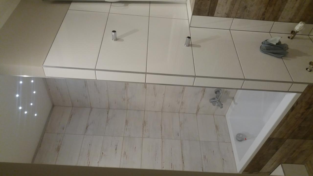 Wykończona łazienka z białymi płytkami na ścianach i podłodze, widoczna wanna, bateria i akcesoria łazienkowe, z dekoracyjnymi płytkami imitującymi drewno na jednej ze ścian.