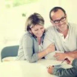 Para w średnim wieku konsultuje dokumenty z doradcą finansowym przy jasnym stole. Uśmiechnięci, zrelaksowani, w tle rozmyte okno.