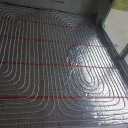 Białe rurki ogrzewania podłogowego ułożone w pętle na srebrnej folii z czerwoną siatką, widok z góry z fragmentem okna po prawej.