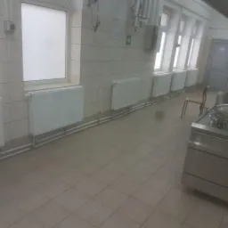 Wnętrze kuchni przemysłowej z grzejnikami pod oknami, widoczne rury instalacji grzewczej i duży stalowy garnek.