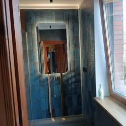 Usługi Glazurnicze Tomasz Mościński - Nowoczesna łazienka z niebieskimi płytkami z wyraźną fakturą, podświetlane lustro z umywalką na stelażu, butelka z płynem do czyszczenia na drewnianym parapecie okna.