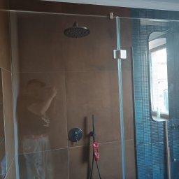 Usługi Glazurnicze Tomasz Mościński - Kabina prysznicowa z deszczownicą i baterią podtynkową na brązowych płytkach, widoczne odbicie osoby robiącej zdjęcie, niebieskie płytki w tle.