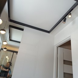 Usługi Glazurnicze Tomasz Mościński - Nowoczesna łazienka z białymi, błyszczącymi panelami ściennymi, czarnymi elementami oświetlenia sufitowego i odbiciem fotografa w lustrze.