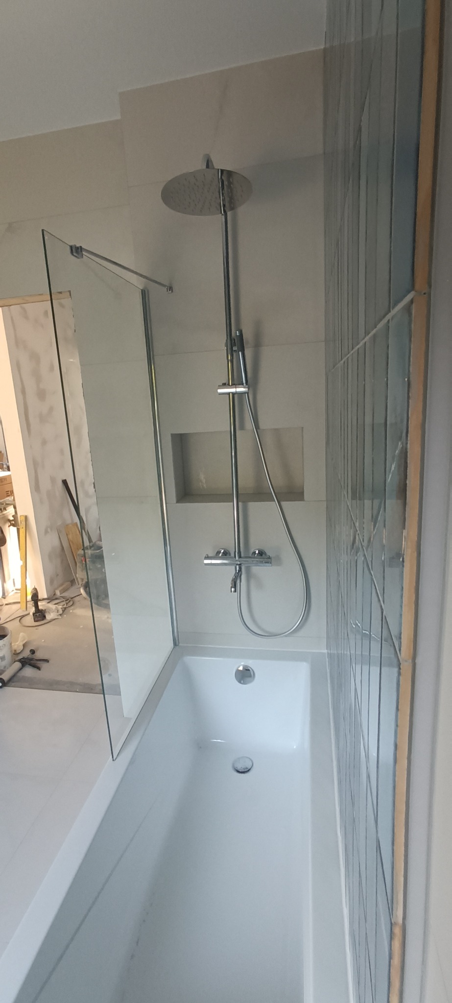 Biała wanna z zamontowaną szklaną ścianką prysznicową i deszczownicą, w tle wnęka w ścianie i fragment ściany wyłożonej wąskimi, pionowymi płytkami w odcieniach błękitu i szarości. W tle widoczne...