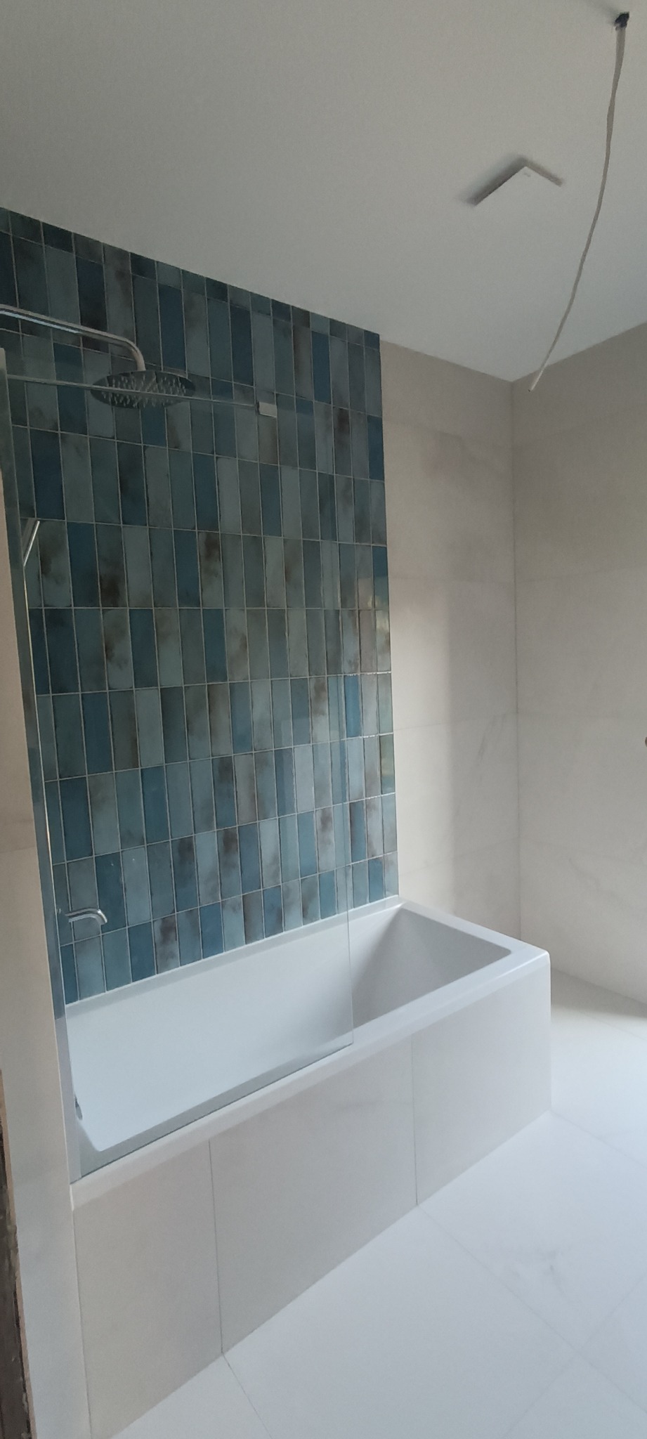 Nowoczesna łazienka z wanną obudowaną białymi płytkami, ściana nad wanną wyłożona pionowymi, niebiesko-szarymi płytkami, szklany parawan nawannowy, deszczownica.